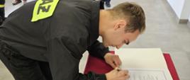 Na zdjęciu w mundurach galowych po lewej strażacy a także pracownicy cywilni. Po prawej strażak składający ślubowanie w ubraniu czarnym koszarowym podpisuje akt ślubowania. Uroczystość ślubowania ma miejsce w sali konferencyjnej.
