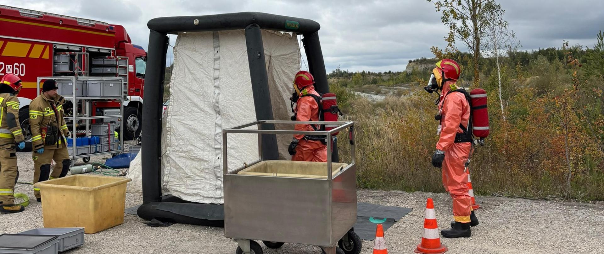 Przy namiocie dekontaminacyjnym PSP dwóch ratowników w pomarańczowych ubraniach ochronnych prowadzi działania. Obok ustawiony jest metalowy wózek na kółkach oraz pojemniki z wodą.