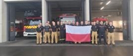 Zdjęcie pamiątkowe. Strażacy trzymają polską flagę stoją przed garażami w których widać trzy strażackie samochody