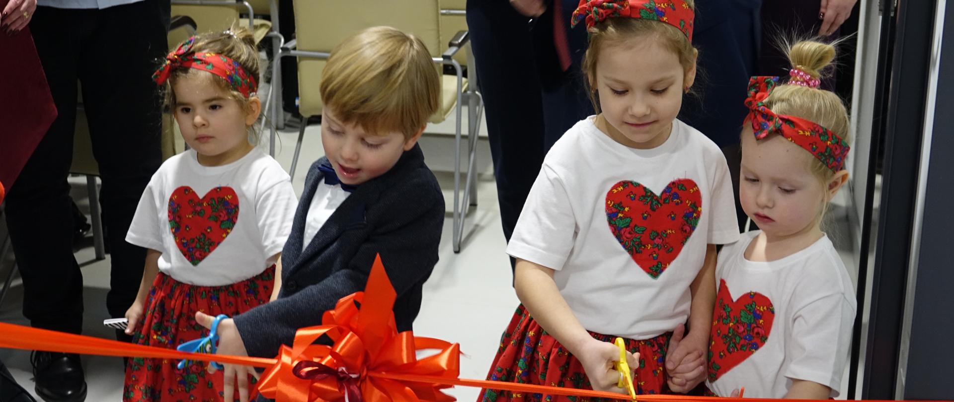 Chłopczyk i trzy dziewczynki otwierają uroczyście żłobek przecinając nożyczkami czerwoną wstęgę.