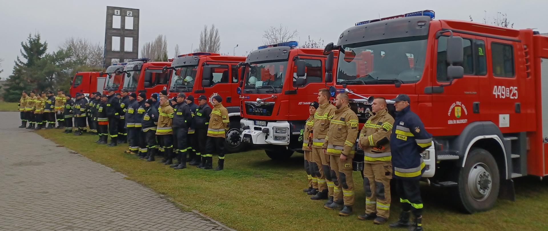Na zdjęciu widać kilkunastu strażaków. Strażacy ubrani są w żółte, żółto-czarne i czarne stroje. Na głowach mają czapki z daszkiem oraz czapki zimowe. Część osób nie ma czapek. Niektórzy strażacy stoją w jednym rzędzie a niektórzy w dwóch rzędach. Za nimi ustawionych jest sześć samochodów strażackich. W tle widać wspinalnie strażacką oraz drzewa. Jest widno.