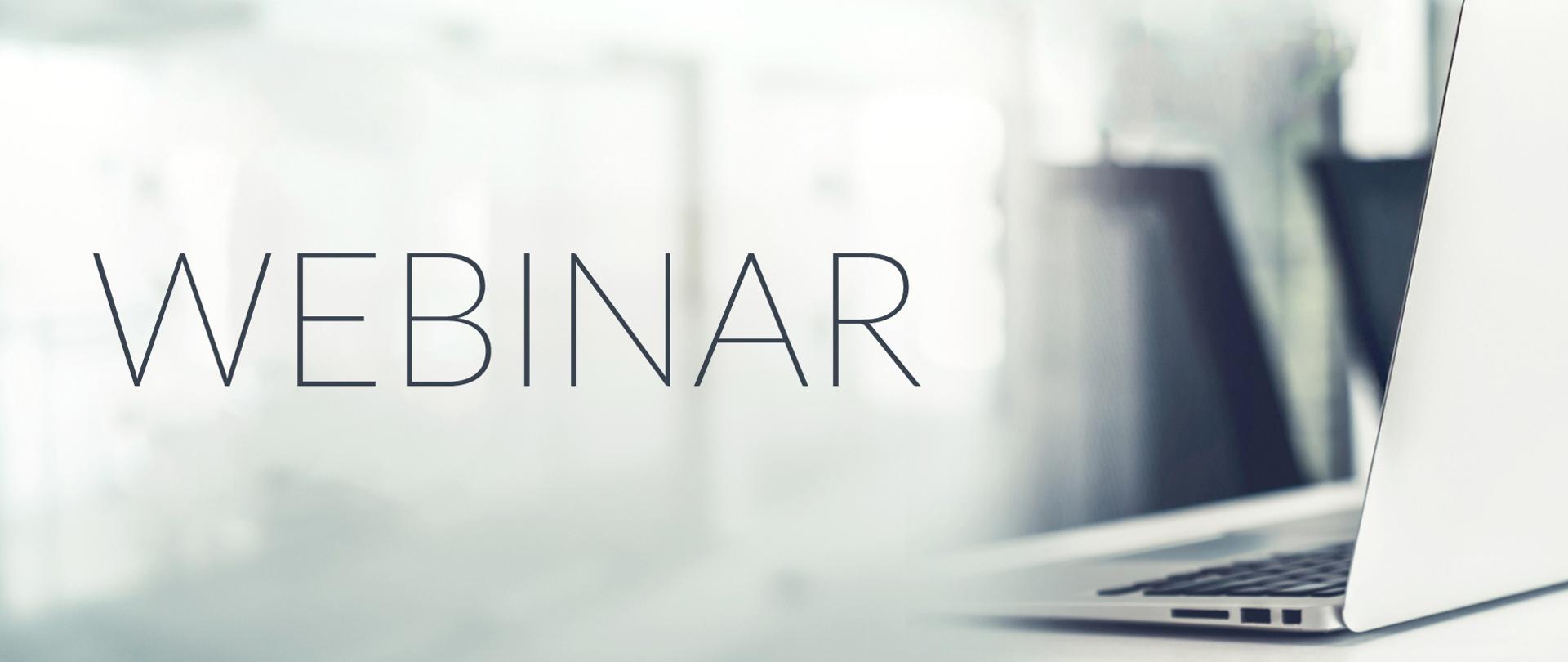 Napis ‘WEBINAR’ na rozmytym tle nowoczesnego biura. Po prawej stronie widoczny jest częściowo otwarty laptop na jasnym biurku.