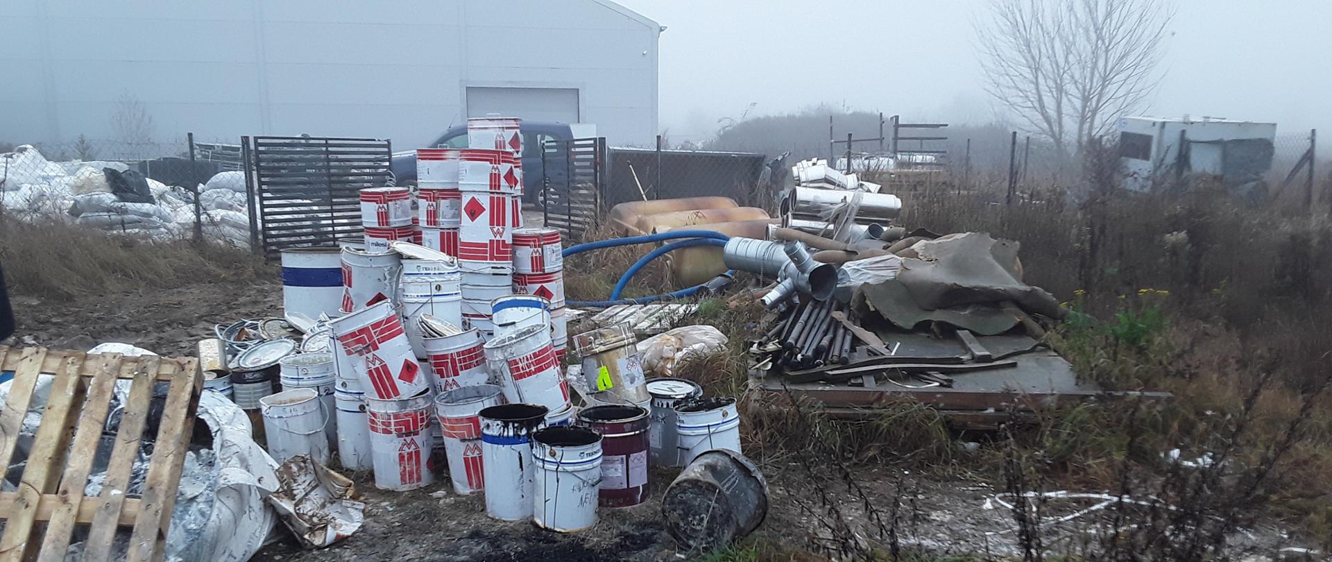 Białe, metalowe pojemniki złożone w nieładzie, jedne na drugich, na otwartym terenie. W tle znajduje się hala.