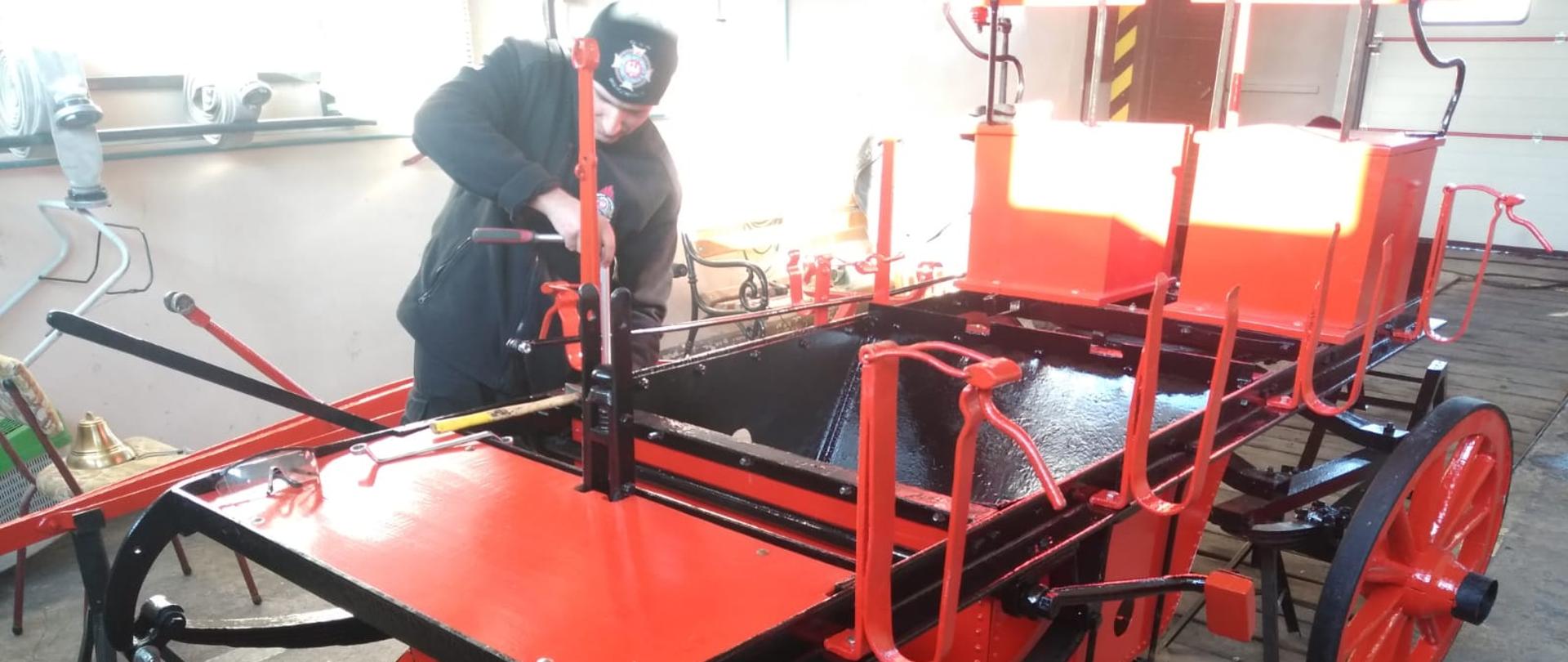 Zdjęcie przedstawia sikawkę konną podczas składania. Jest ona pomalowana na czerwono i czarno. Jeden ze strażaków maluje ją.