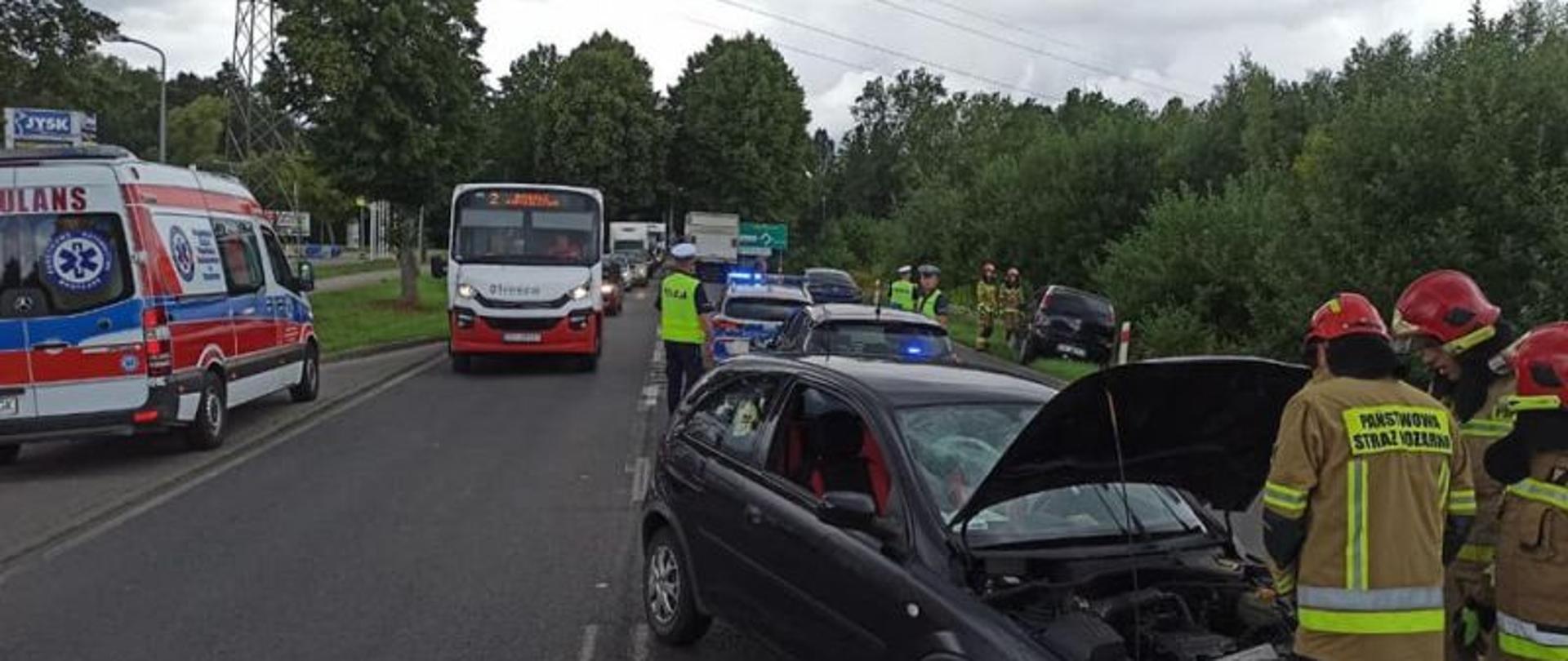 Zdjęcie wykonane w dzień. Zdjęcie przedstawia miejsce zderzenia pojazdów. Na drodze panuje duży ruch. Od prawej grupa strażaków odłączająca akumulator od rozbitego pojazdu. Na lewo inne pojazdy mijające miejsce zdarzenia. W tle niebieskie niebo z dużymi chmurami.
