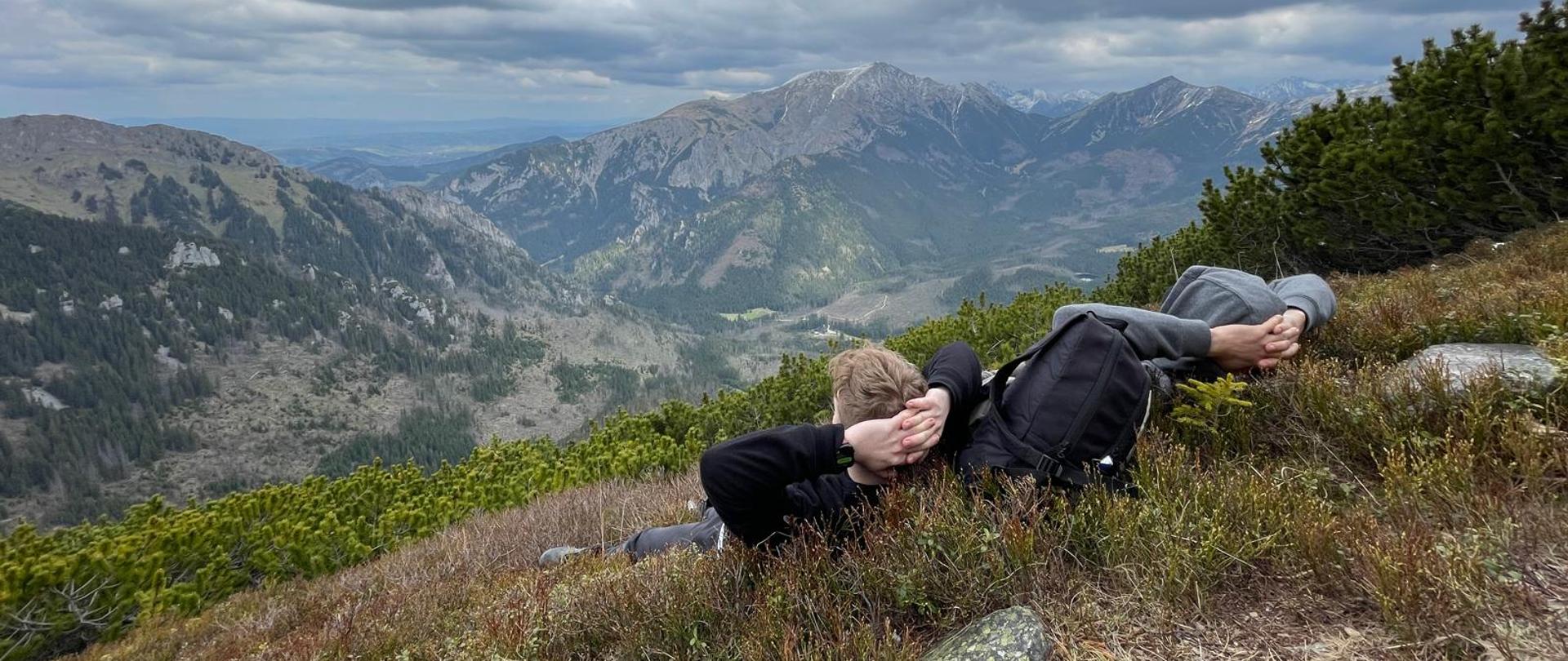 Leżące na plecach, na łagodnym zboczu góry dwie, oglądające górskie widoki, osoby. Po horyzont rozciągają się górskie szczyty i pasma. Niebo poszyte jest gęstymi, ciemnymi chmurami, zbocza częściowo roślinnością i łatami śniegu. 