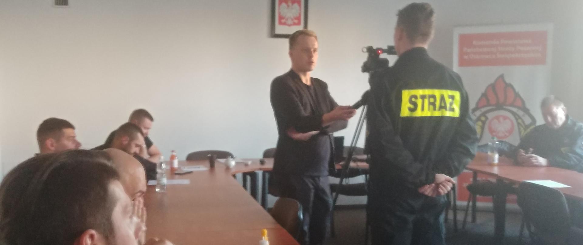 Na zdjęciu w pomieszczeniu budynku Komendy powiatowej PSP w Ostrowcu Św. odbywa się szkolenie dla dowódców OSP z terenu OSP. Na pierwszym planie widać druhów, którzy siedząc przyglądają się jak jednej ze szkolących udziela wywiadu przed kamerą.