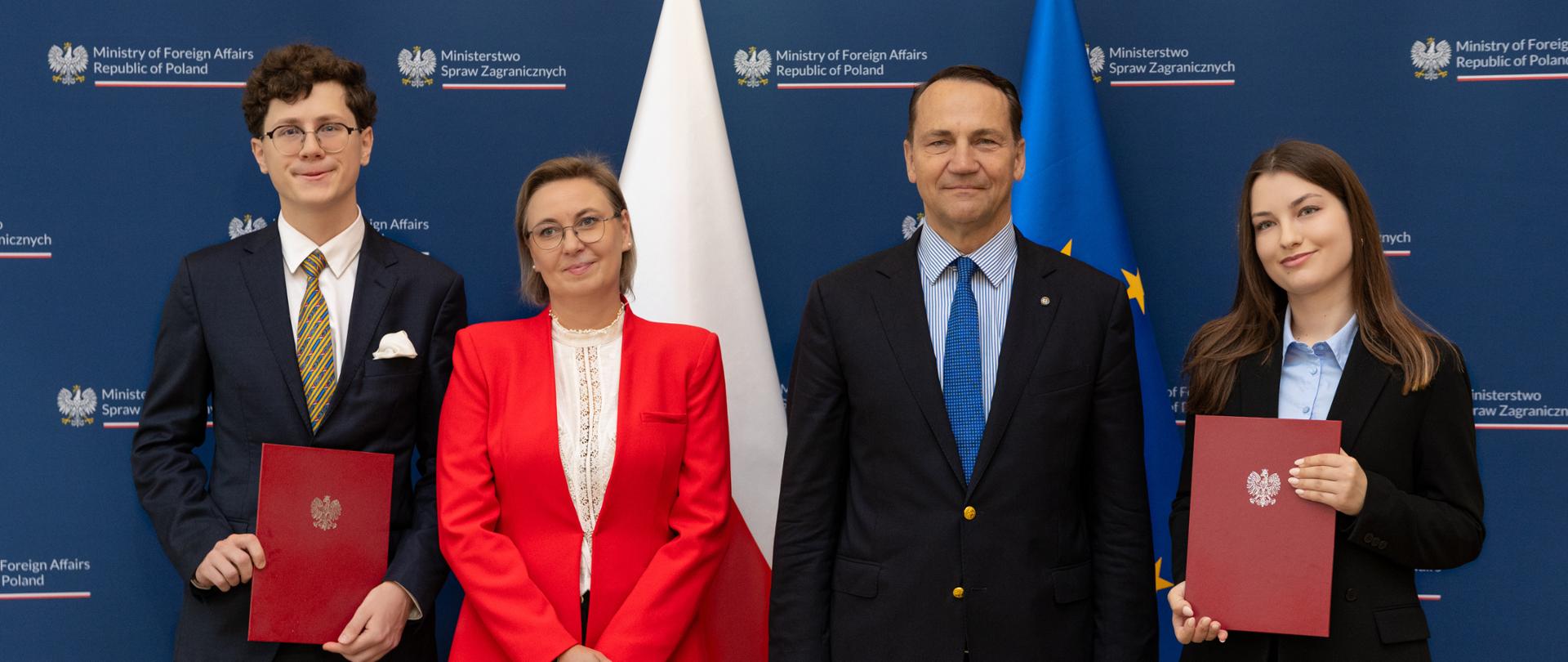 Na zdjęciu są 4 osoby. Stoją obok siebie i patrzą na wprost. Dwie osoby trzymają czerwone teczki. Za nimi są flagi: Polski i UE. 