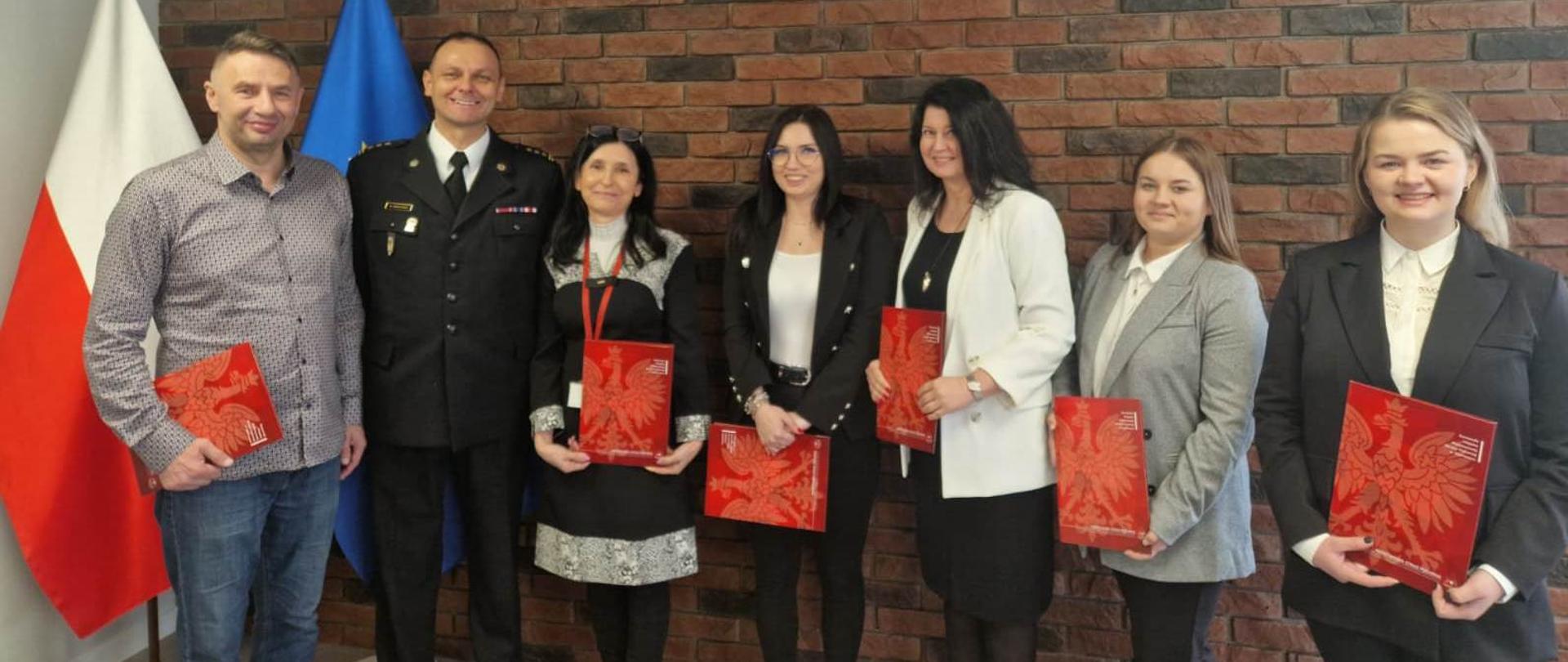Na zdjęciu widoczna jest grupa sześciu osób stojących w siedzibie jednostki na tle ceglanej ściany. Za nimi znajdują się flagi Polski i Unii Europejskiej. Jedna z osób ubrana jest w mundur strażacki, pozostali w stroje cywilne o charakterze formalnym. Część osób trzyma w rękach czerwone teczki z wizerunkiem orła. Wszyscy stoją obok siebie i pozują do wspólnego, pamiątkowego zdjęcia.
