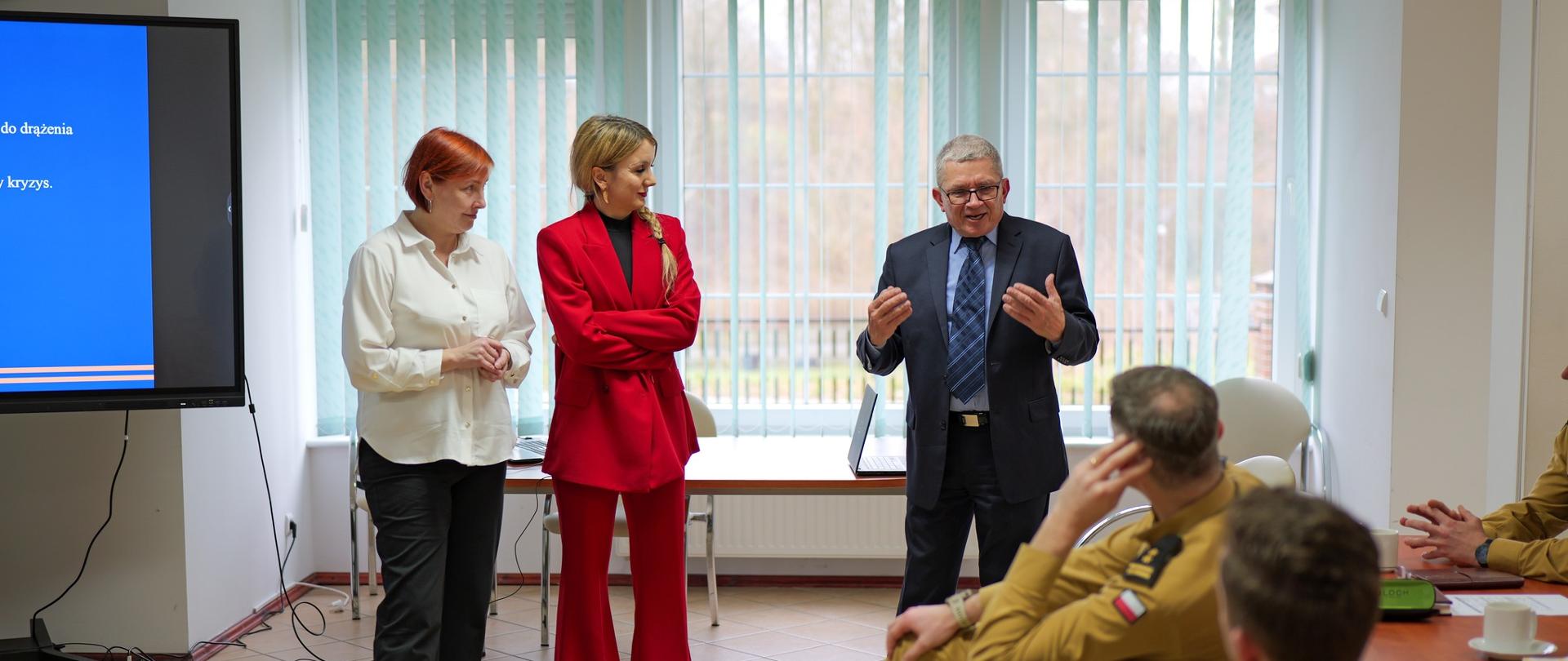 Trzy osoby – dwie kobiety i jeden mężczyzna (prowadzący szkolenie) – stoją przed grupą strażaków. Kobieta po lewej stronie ma białą koszulę, środkowa nosi czerwony garnitur, a mężczyzna w garniturze gestykuluje, tłumacząc temat. Za nimi ekran z prezentacją.
