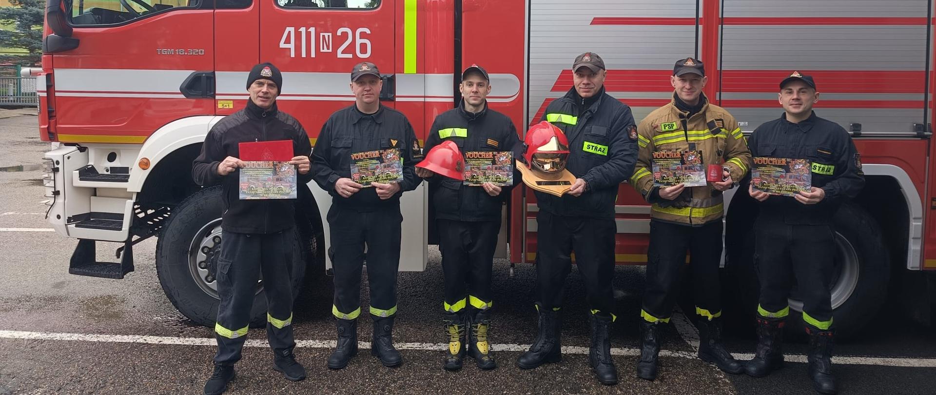 Na zdjęciu przy samochodzie strażackim stoją strażacy z upominkami , które wystawia na licytacje WOŚ.