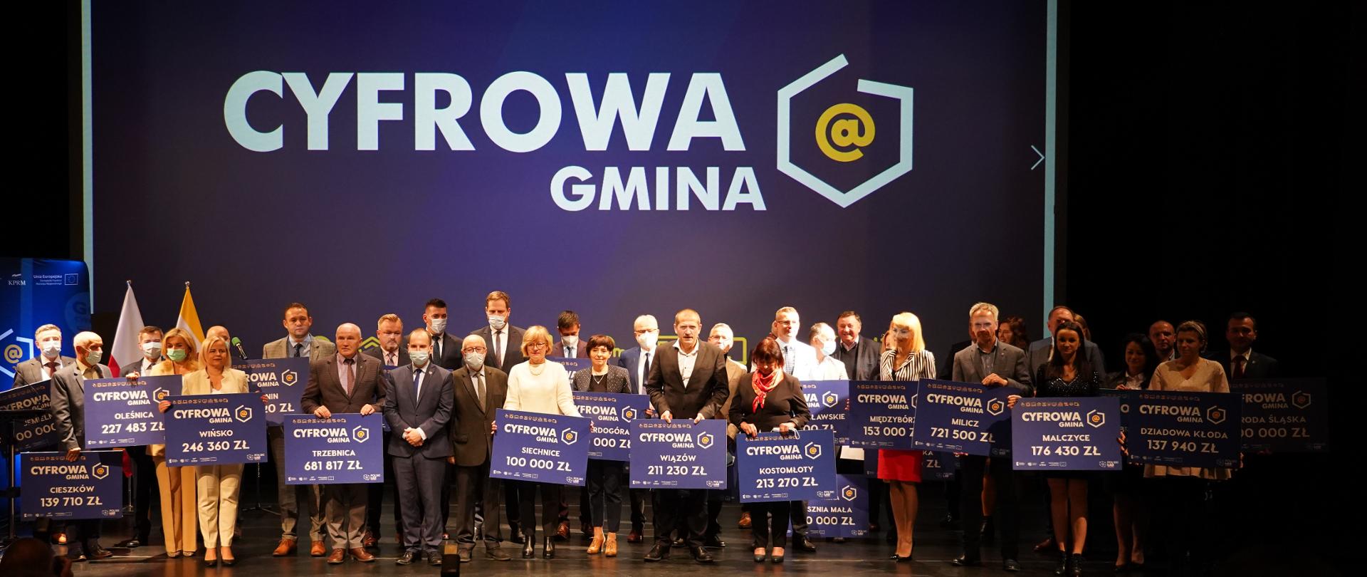 Ministrowie Janusz Cieszyński i Adam Andruszkiewicz w Oleśnicy wśród samorządowców trzymających symboliczne czeki z kwotami wsparcia, które mogą otrzymać z programu Cyfrowa Gmina.