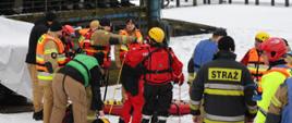 Grupa ratowników strażaków w kolorowych kombinezonach i kaskach podczas ćwiczeń przy zamarzniętym zbiorniku wodnym. Na pierwszym planie przygotowują sprzęt i linę ratunkową przy brzegu, a w tle na pomoście stoi grupa obserwujących osób. Widać śnieg, lód i zimową scenerię