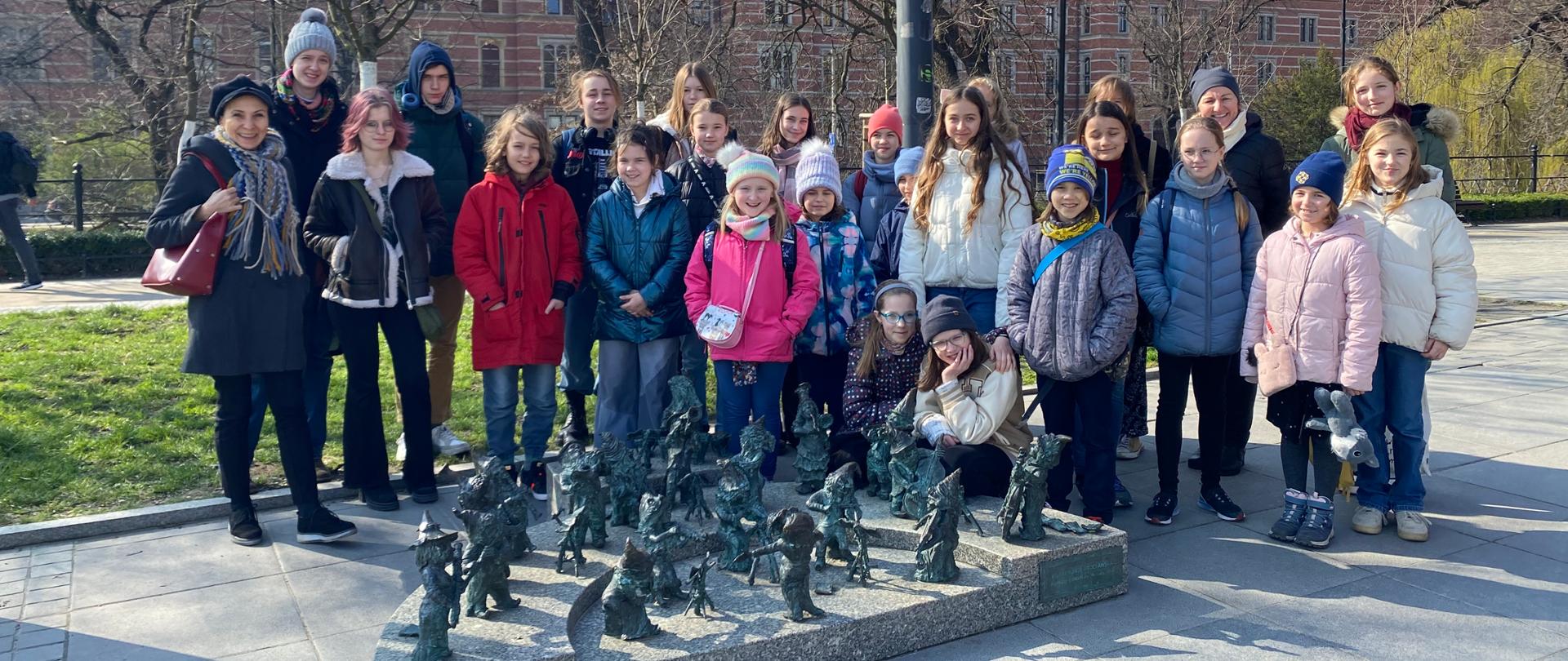 Grupa młodzieży wraz z nauczycialami. Grupa stoi na zewnątrz Narodowego Forum Muzyki przy figurach krasnoludkowej orkiestry symfonicznej , w tle budynek wrocławskiego sądu (zbudowany z czerwonej cegły).