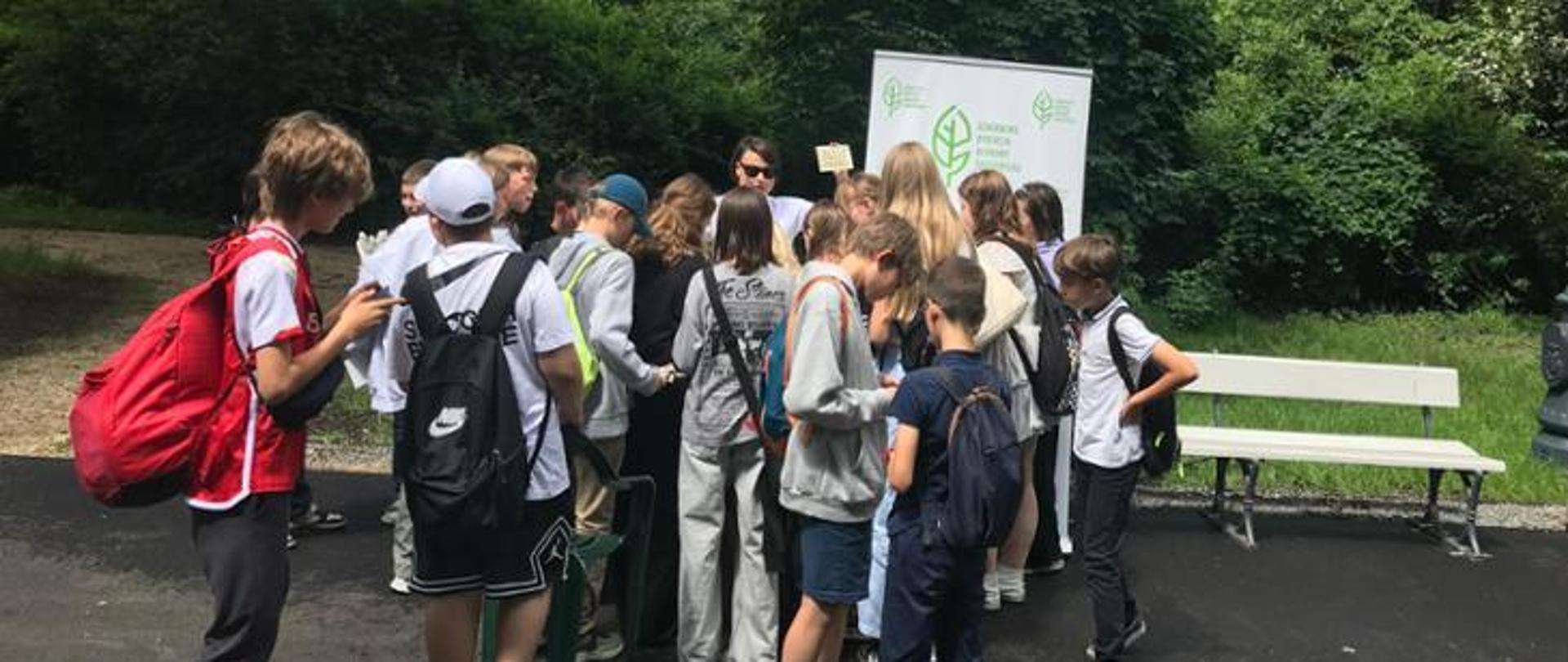 Na stoisku informacyjnym Generalnej Dyrekcji Ochrony Środowiska kobieta opowiada o roślinach miododajnych, otacza ją grupa nastolatków, w tle widać ściankę z logo urzędu, oraz zieleń kwitnących drzew.