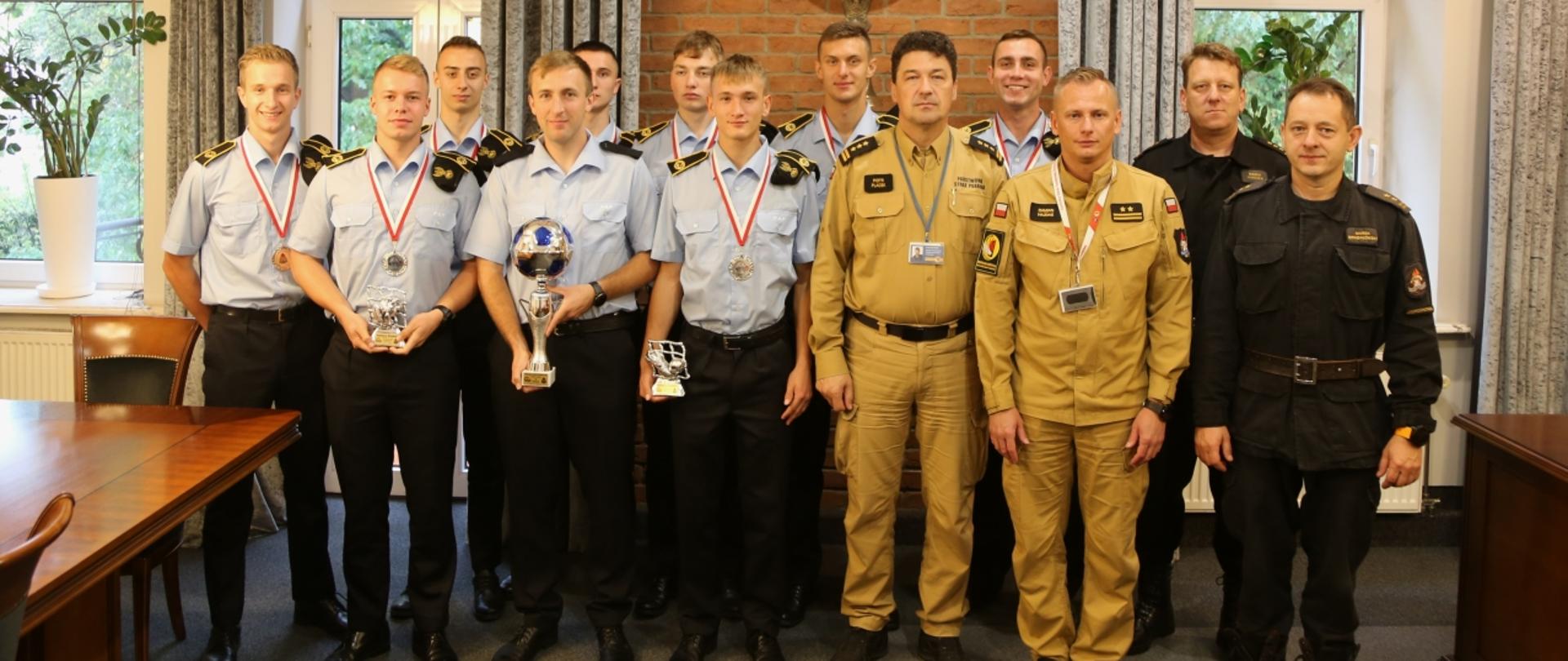 Grupowe zdjęcie Komendanta Centralnej Szkoły PSP, Zastępcy Komendanta CS PSP, naczelników wydziału kompanii szkolnych i zabezpieczeń obiektów oraz wydziału nauczania w towarzystwie reprezentacji CS PSP w piłce nożnej 6-osobowej (9 osób). Zawodnicy mają na szyi zdobyte srebrne medale oraz trzymają zdobyty puchar oraz statuetki za wyróżnienia indywidualne