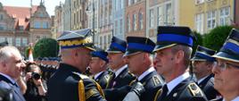 Strażak w mundurze galowym przypina odznaczenie strażakowi obok w rzędzie stoją inni strażacy oraz obok mężczyzna w garniturze i fotograf trzymający aparat.