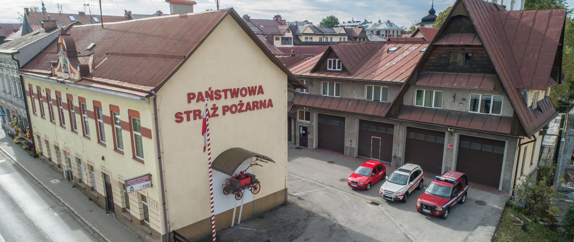 Na zdjęciu znajdują się budynki komendy powiatowej psp w Nowym Targu przed bramami ustawione są trzy samochody operacyjne, na bocznej ścianie jednego z budynków zawieszona jest zabytkowa sikawka