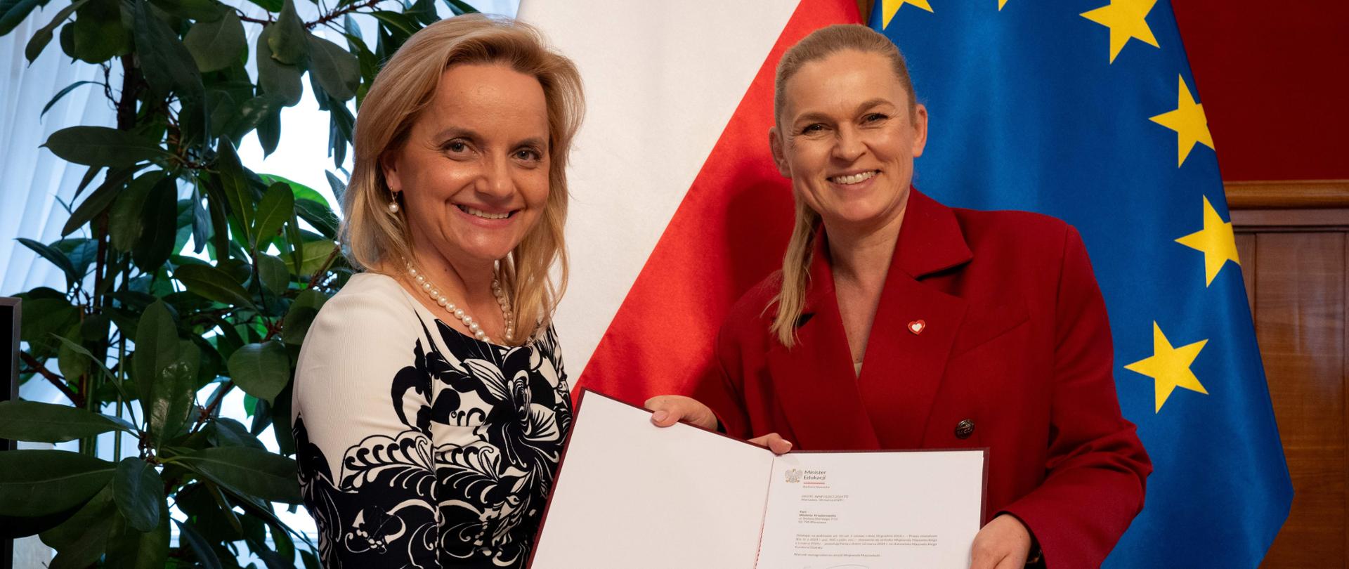 Minister Nowacka stoi obok Wioletty Krzyżanowskiej. Obie panie w ręce trzymają otwarty dokument powołania. W tle flagi Unii Europejskiej i Polski.
