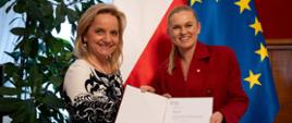 Minister Nowacka stoi obok Wioletty Krzyżanowskiej. Obie panie w ręce trzymają otwarty dokument powołania. W tle flagi Unii Europejskiej i Polski.