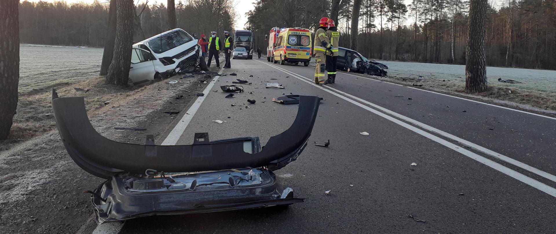 Na zdjęciu widać pas drogowy, na którym na pierwszym planie leży urwany zderzak pojazdu. W dalszej części w rowach po przeciwnych stronach widać dwa rozbite pojazdy. Na drodze stoi karetka pogotowia i samochód pożarniczy dwóch strażaków i 2 policjantów.
