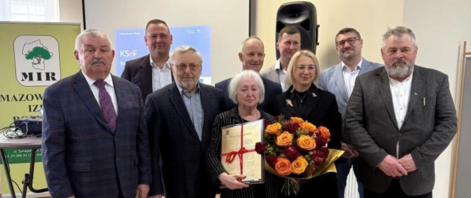 Grupa osób stojących do zdjęcia w sali konferencyjnej; jedna z osób trzyma dyplom i bukiet kwiatów, co sugeruje uroczyste wręczenie wyróżnienia.