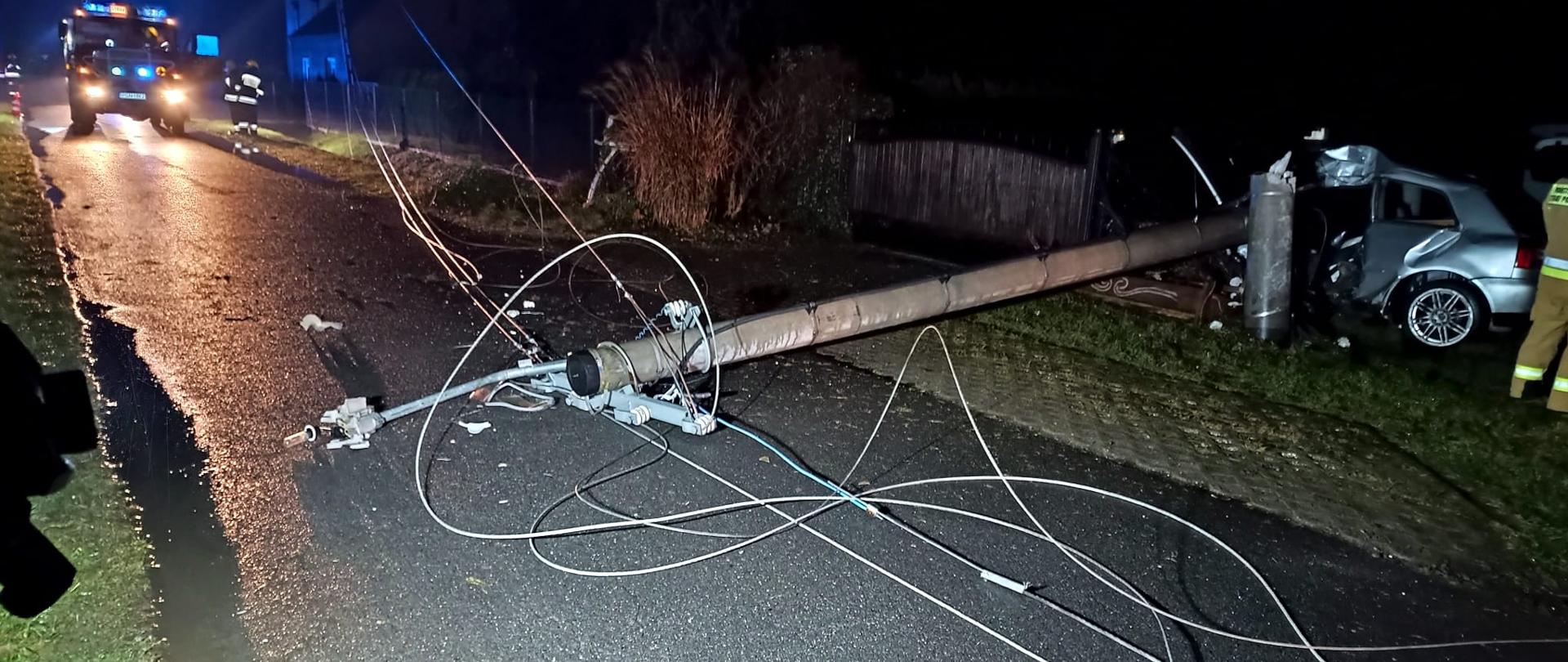 Jest pora nocna, na drodze leży ścięty słup energetyczny oraz linie energetyczne tarasujące całkowicie przejazd drogą. Z lewej strony zdjęcia widać bok uszkodzonego pojazdu. W tle na drodze stoi samochód pożarniczy.