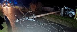 Jest pora nocna, na drodze leży ścięty słup energetyczny oraz linie energetyczne tarasujące całkowicie przejazd drogą. Z lewej strony zdjęcia widać bok uszkodzonego pojazdu. W tle na drodze stoi samochód pożarniczy.