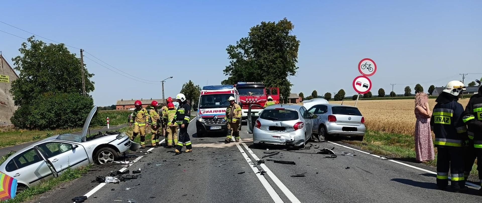 Zdjęcie przedstawia trzy rozbite samochody (wszystkie koloru srebrnego) jeden pojazd znajduje się w rowie, dwa pozostałe stoją na drodze. Przy pojazdach stoją strażacy i policjanci oraz wóz strażacki i karetka.