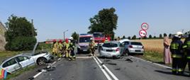 Zdjęcie przedstawia trzy rozbite samochody (wszystkie koloru srebrnego) jeden pojazd znajduje się w rowie, dwa pozostałe stoją na drodze. Przy pojazdach stoją strażacy i policjanci oraz wóz strażacki i karetka.