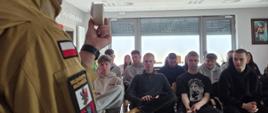 Na zdjęciu przedstawiono zajęcia edukacyjne prowadzone przez strażaka w sali szkoleniowej. Instruktor, ubrany w mundur, stoi przodem do grupy i prezentuje niewielkie urządzenie – prawdopodobnie czujnik dymu lub element systemu bezpieczeństwa pożarowego.
Przed nim siedzi grupa młodych osób, które uważnie słuchają i obserwują prezentację. Uczestnicy zajęć zajmują miejsca na krzesłach ustawionych w rzędach. Całość przedstawia spotkanie o charakterze edukacyjnym, mające na celu zwiększenie świadomości w zakresie bezpieczeństwa oraz ochrony przeciwpożarowej.