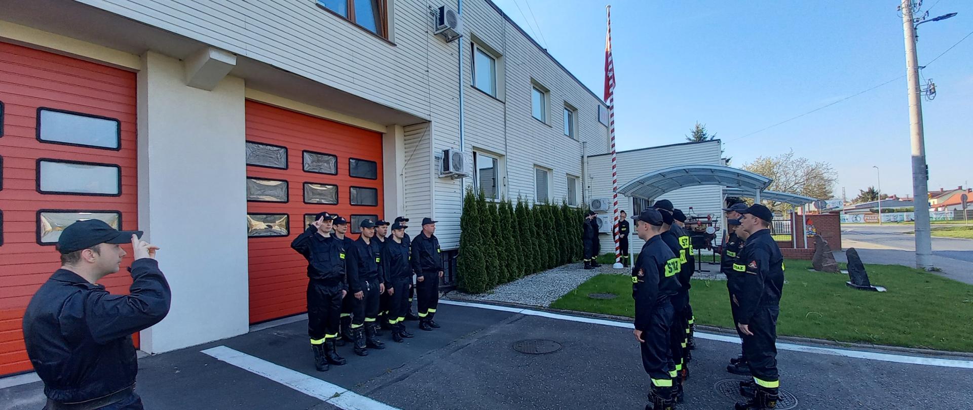 Zdjęcie przedstawia uroczystą zmianę służby Jednostki Ratowniczo Gaśniczej przed budynkiem KP PSP w Zduńskiej Woli podczas podniesienia flagi państwowej RP. W zmianie służby uczestniczą funkcjonariusze w umundurowaniu koszarowym, zmiany służbowej III - zdającej, zmiany służbowej I - przyjmującej, Dowódca JRG oraz poczet flagowy.