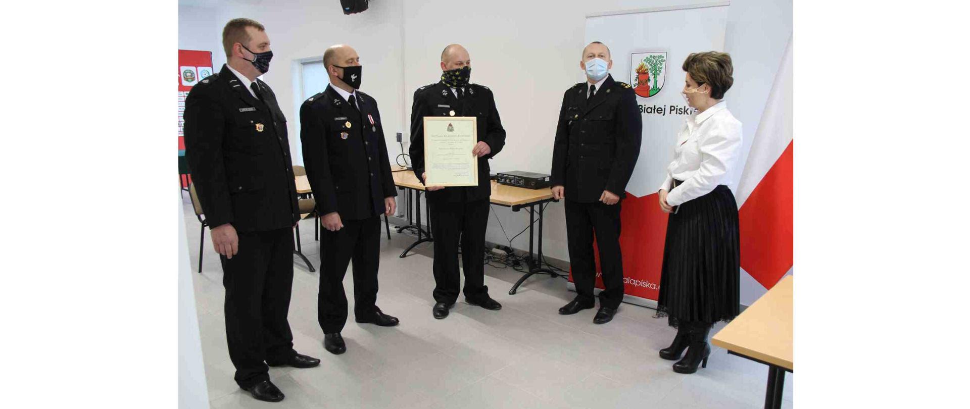 Burmistrz Białej Piskiej oraz czterech strażaków stojących w pomieszczeniu. jeden z nich trzyma decyzję Komendanta Głównego PSP o włączeniu jednostki OSP Skarżyn do Krajowego Systemu Ratowniczo-Gaśniczego.