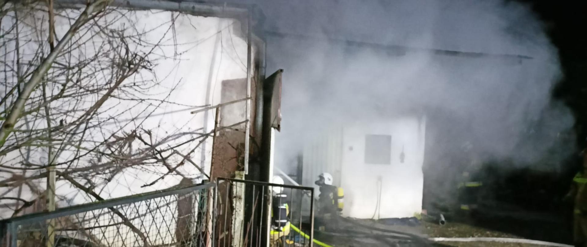 Widać wydobywający się dym z budynku oraz strażacy, którzy gaszą pożar