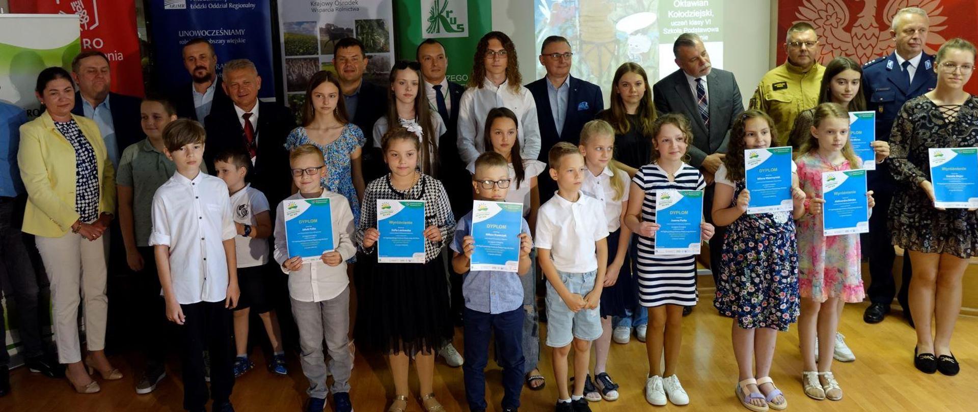 Grupa dzieci z dyplomami w rękach a z tyłu stoi grupa dorosłych mężczyzn i kobiet.