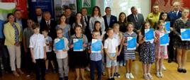 Grupa dzieci z dyplomami w rękach a z tyłu stoi grupa dorosłych mężczyzn i kobiet.