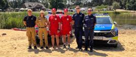 Przedstawiciele służb wspólnie pozują do zdjęcia, które zostało wykonane na plaży na tle samochodów służbowych.