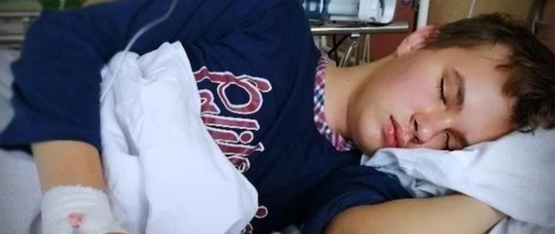 Mateusz leży lewym bokiem na łóżku szpitalnym, chłopiec ma zamknięte oczy, w tle aparatura szpitalna