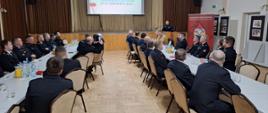 Strażacy ubrani w granatowe mundury galowe, oraz pisakowe mundury siedzą przy stolikach, w tyle rolap z logiem Państwowej Straży Pożarnej na tylnej ścianie kotary oraz ekran z wyświetloną prezentacją. Przy mównicy stoi prezes ubrany w granatowy polar, przed sobą ma mikrofon