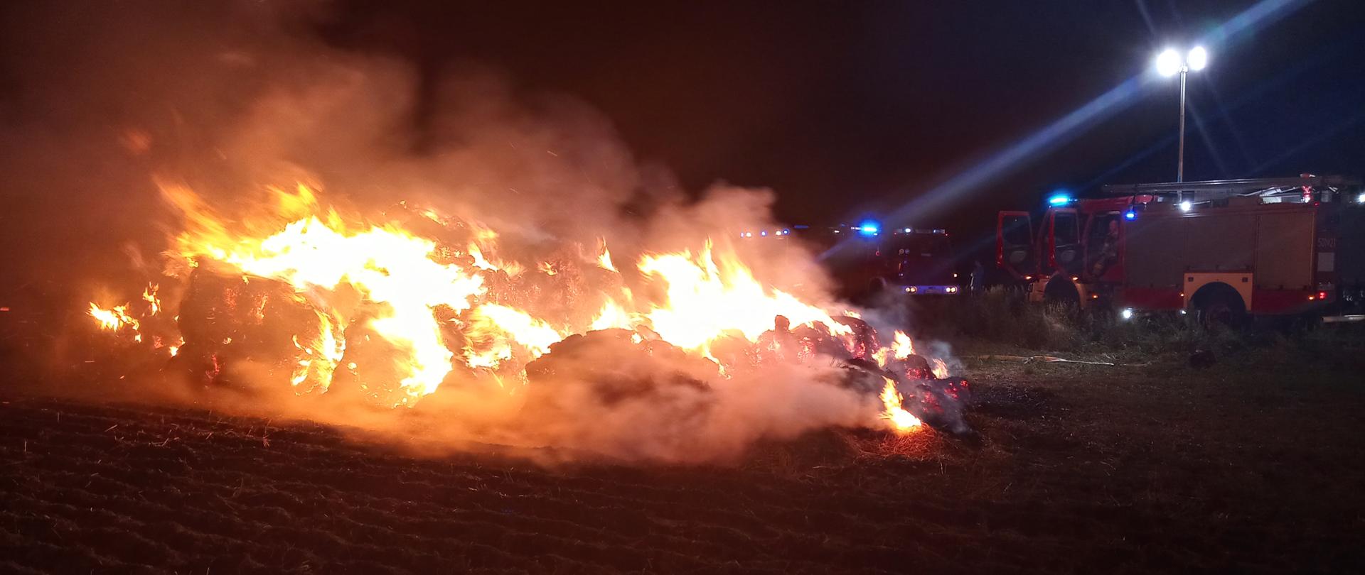 Pali sie stóg balotów słomy. W tle stoją pojazdy straży pożarnej.