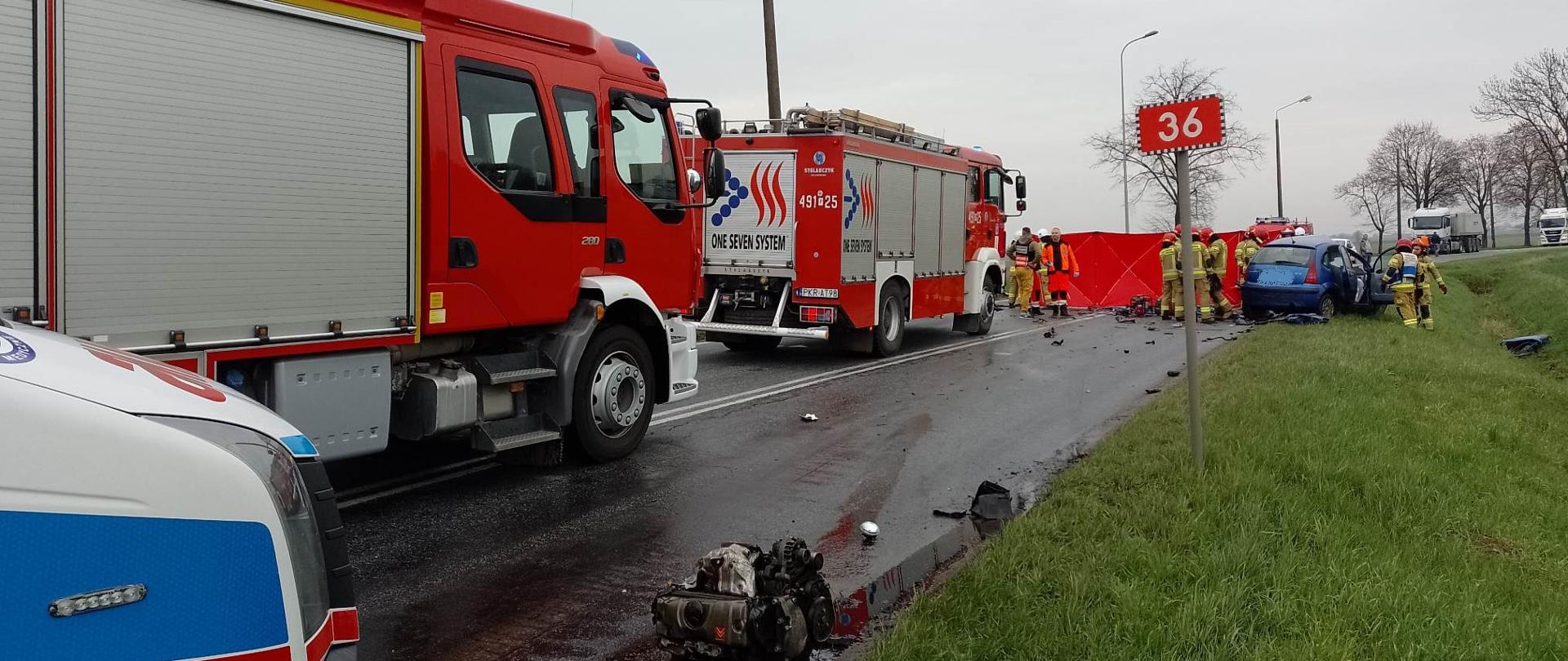 Zdjęcie przedstawia samochody służb ratowniczych tj. Straży Pożarnej i Karetkę pogotowia stojące na drodze oraz samochód osobowy koloru niebieskiego, który brał udział w zdarzeniu. Wokół rozbitego pojazdu wykonuję swoje czynności strażacy i ratownicy medyczni. Zdjęcie przedstawia również leżący na poboczu drogi silnik z uszkodzonego pojazdu i znak drogowy z numerem drogi "36".