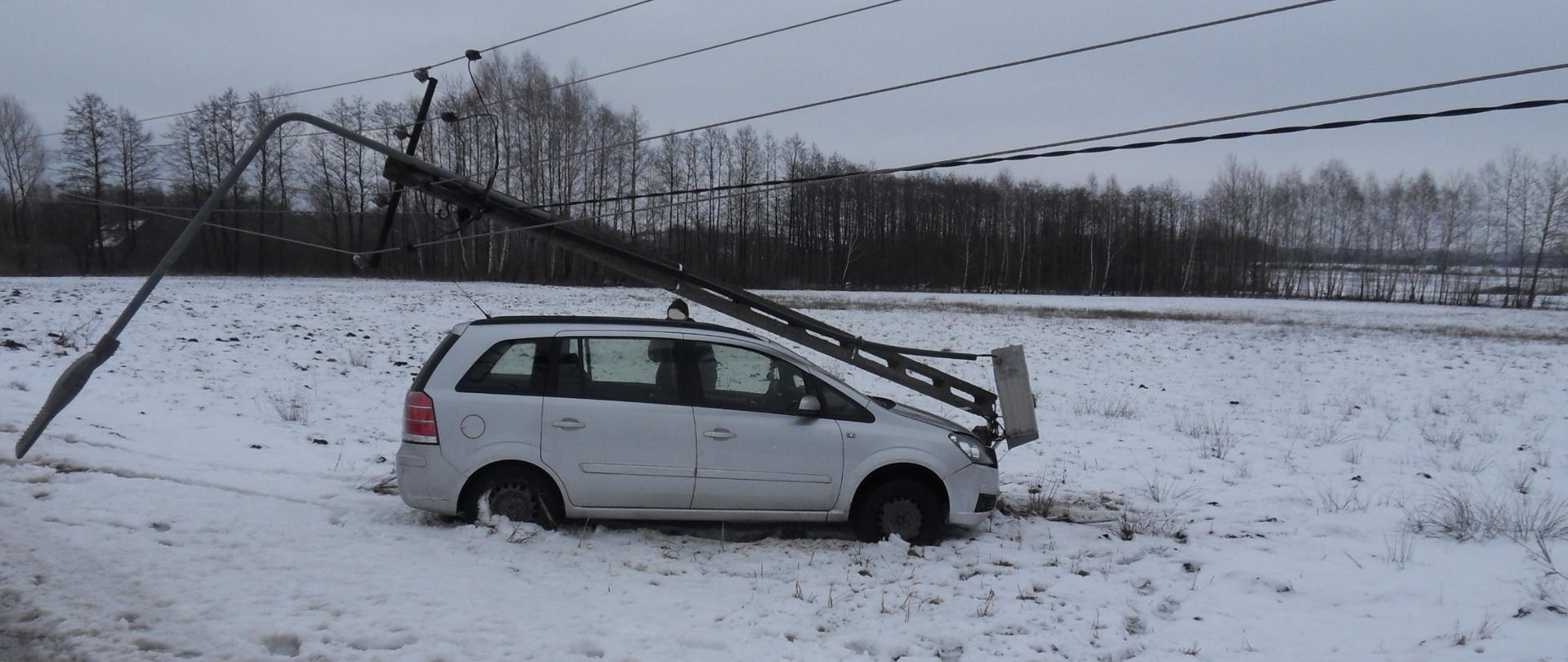 Samochód osobowy w kolorze srebrnym, który przodem uderzył w słup elektryczny, nad pojazdem wisi złamany słup elektryczny. Samochód stoi na ośnieżonej łące. 
