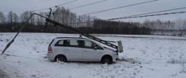 Samochód osobowy w kolorze srebrnym, który przodem uderzył w słup elektryczny, nad pojazdem wisi złamany słup elektryczny. Samochód stoi na ośnieżonej łące. 