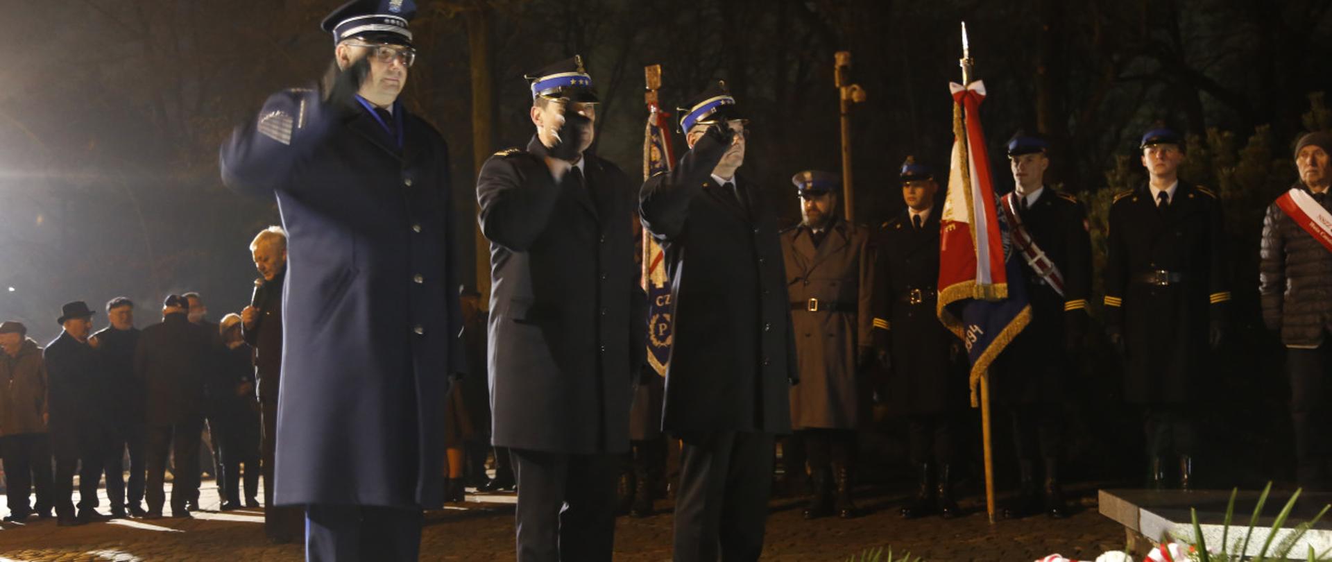 Oficer policji, Komendant Centralnej Szkoły PSP oraz Zastępca Komendanta Centralnej Szkoły PSP oddają honor salutując stojąc przed pomnikiem przed którym leżą wieńce, w głębi widoczny poczet sztandarowy Centralnej Szkoły PSP