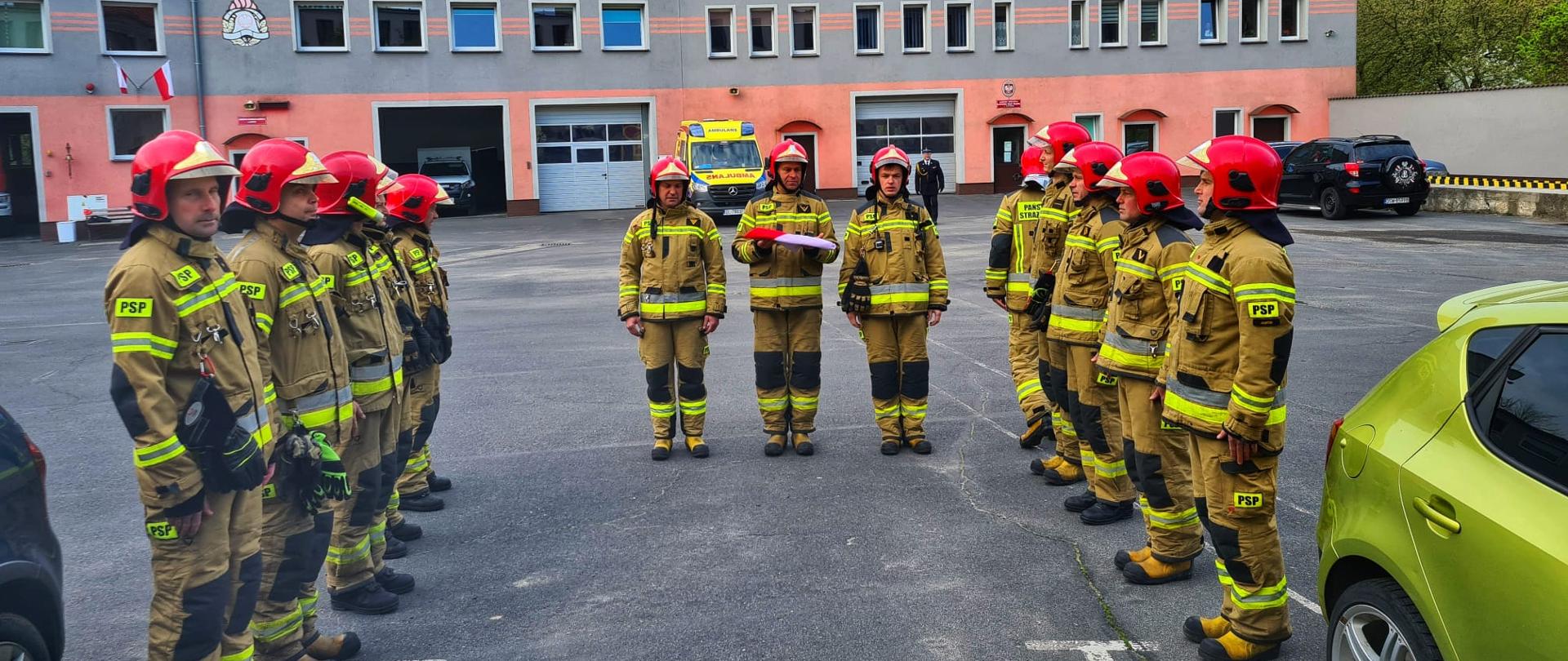 Obraz przedstawia strażaków Komendy Powiatowej Państwowej Straży Pożarnej w Jaworze podczas uroczystej zbiórki z okazji Dnia Flagi. Strażacy w umundurowaniu specjalnym stoją naprzeciw siebie w dwóch szeregach. W tle budynek komendy.