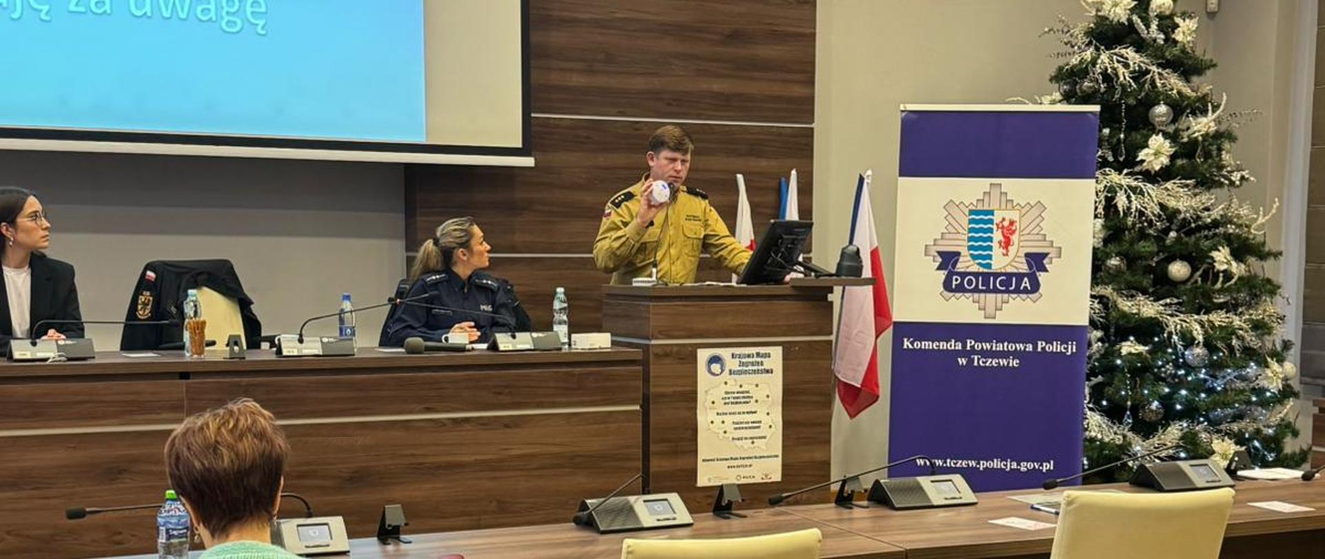 Strażak prezentuje czujkę czadu na mównicy w urzędzie miasta. obok policjanci i choinka.
