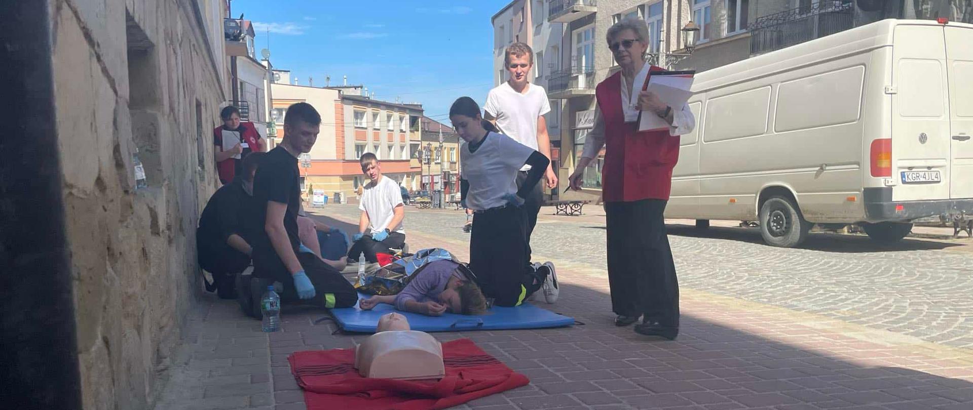 Ćwiczenia z ratownictwa przed kamienicą. Młodzież w rękawiczkach jednorazowych opiekuje się pozorantem leżącym w pozycji bocznej bezpiecznej.