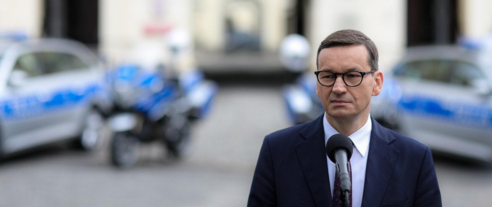 Premier Mateusz Morawiecki na tle samochodów policji.