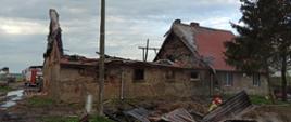 Zdjęcie przedstawia od lewej: budynek gospodarczy po pożarze ze spalonym dachem. Obok budynku stoi wóz strażacki. Obok budynku gospodarczego budynek mieszkalny z nadpalonym dachem. Z przodu leżą metalowe pozostałości dachu