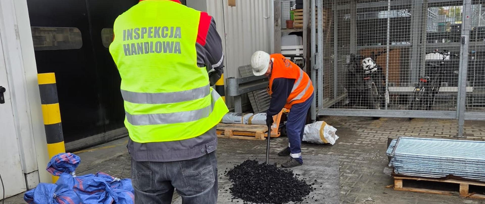 Na pierwszym planie niebieskie duże worki związane u góry, na środkowym planie stoi mężczyzna w kamizelce z napisem Inspekcja Handlowa, na dalszym planie mężczyzna z łopatą nabiera węgiel, który leży na plandece. 